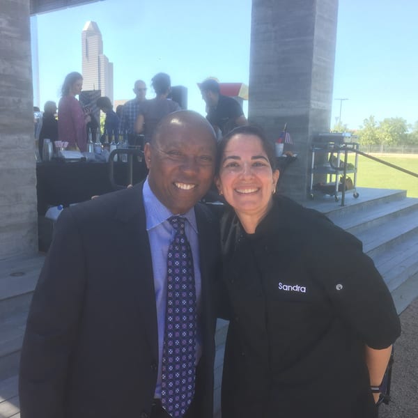 A man in a suit and a woman in a chef's coat smile, with a city skyline in the background.
