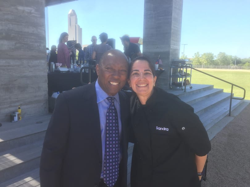 A man in a suit and a woman in a chef's coat smile, with a city skyline in the background.