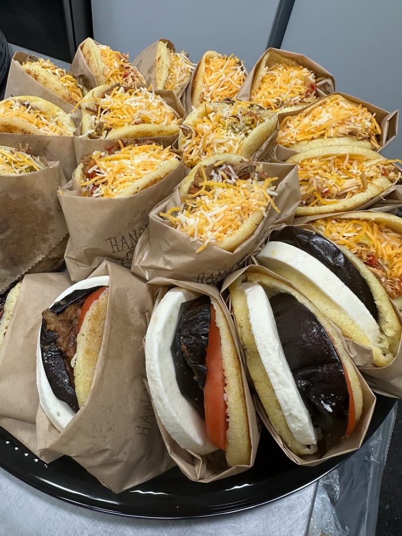 A tray of various arepas, some with shredded cheese, others with eggplant, tomato, and cheese.