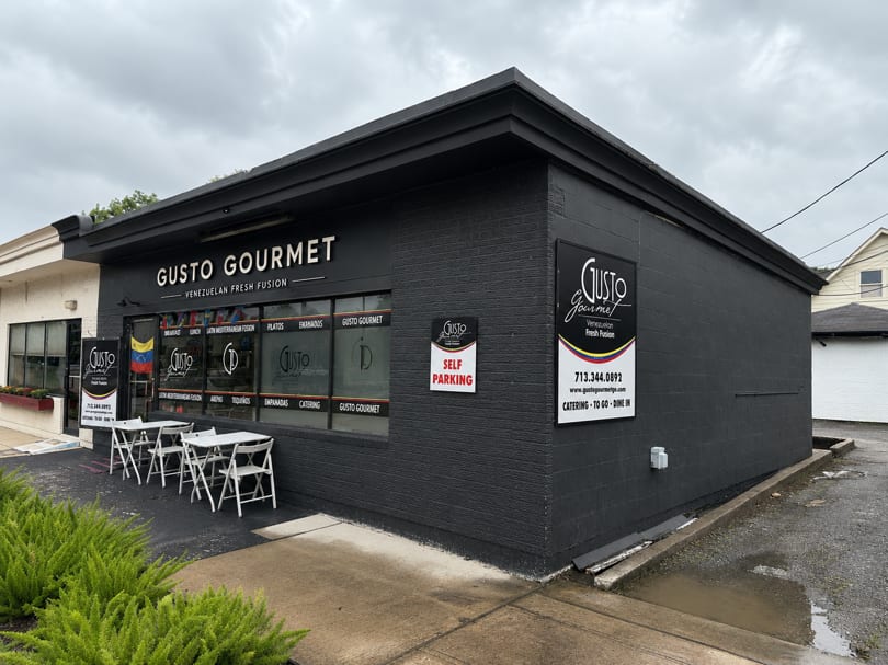 Exterior of Gusto Gourmet, a Venezuelan Fresh Fusion restaurant, with outdoor seating and dark gray facade.