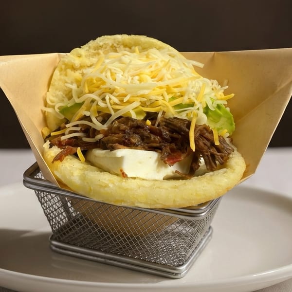 Arepa with shredded beef, white cheese, avocado, and shredded yellow cheese in a metal basket.