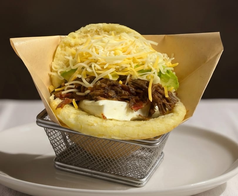Arepa with shredded beef, white cheese, avocado, and shredded yellow cheese in a metal basket.