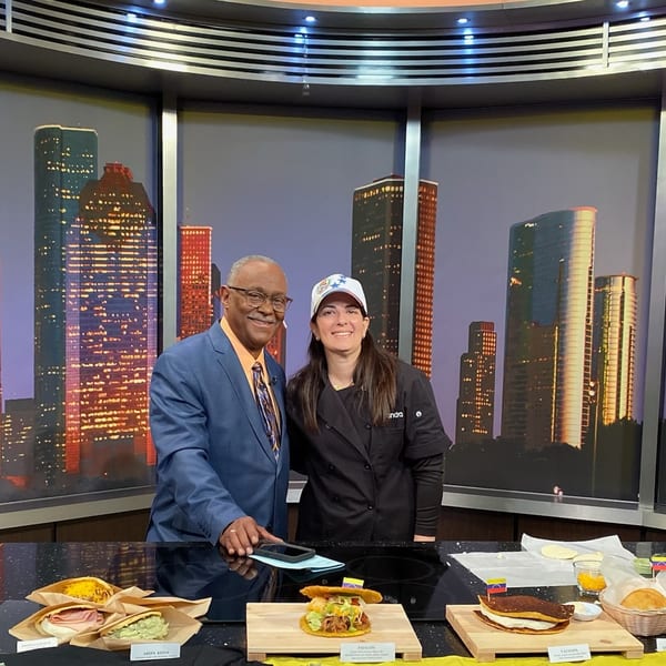 A chef and a man in a suit stand behind a counter with various Venezuelan dishes and ingredients.