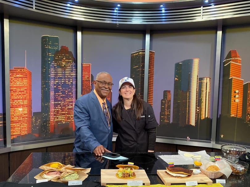 A chef and a man in a suit stand behind a counter with various Venezuelan dishes and ingredients.