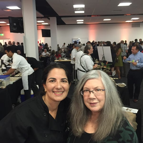Two women smiling at the camera in a crowded event hall with chefs and guests.