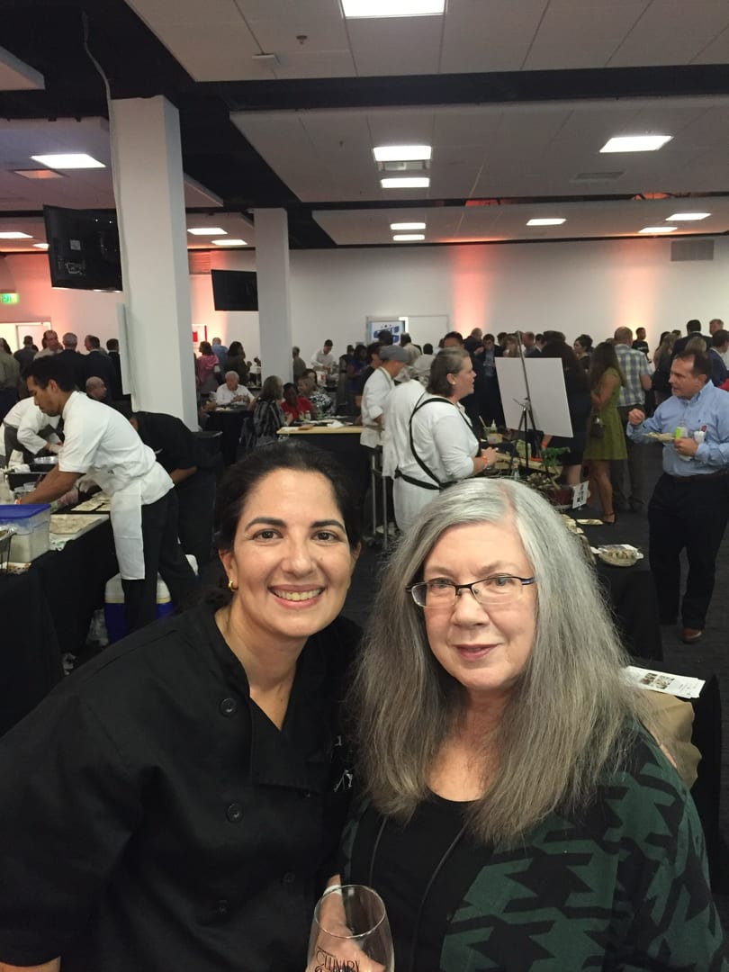 Two women smiling at the camera in a crowded event hall with chefs and guests.