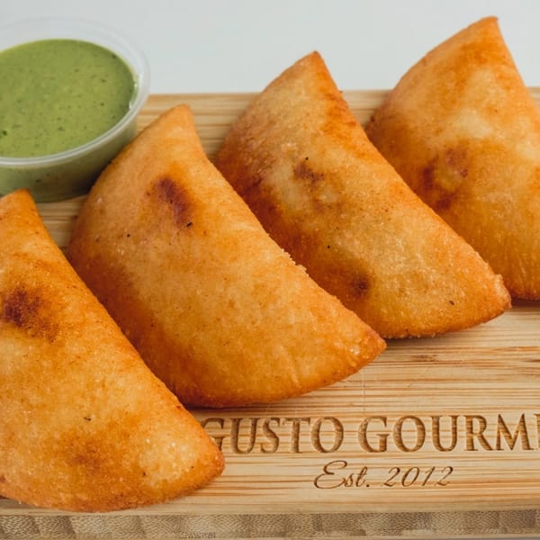Four golden-brown empanadas and a small cup of green sauce on a wooden board from Gusto Gourmet.