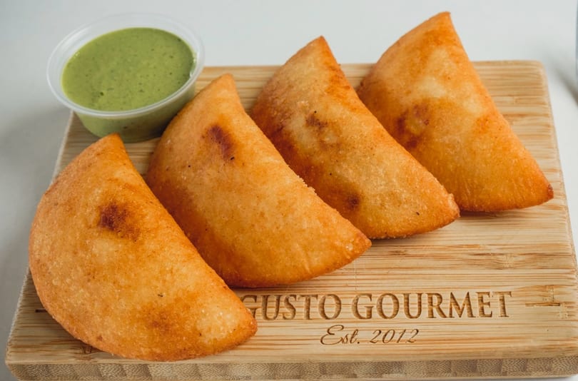 Four golden-brown empanadas and a small cup of green sauce on a wooden board from Gusto Gourmet.